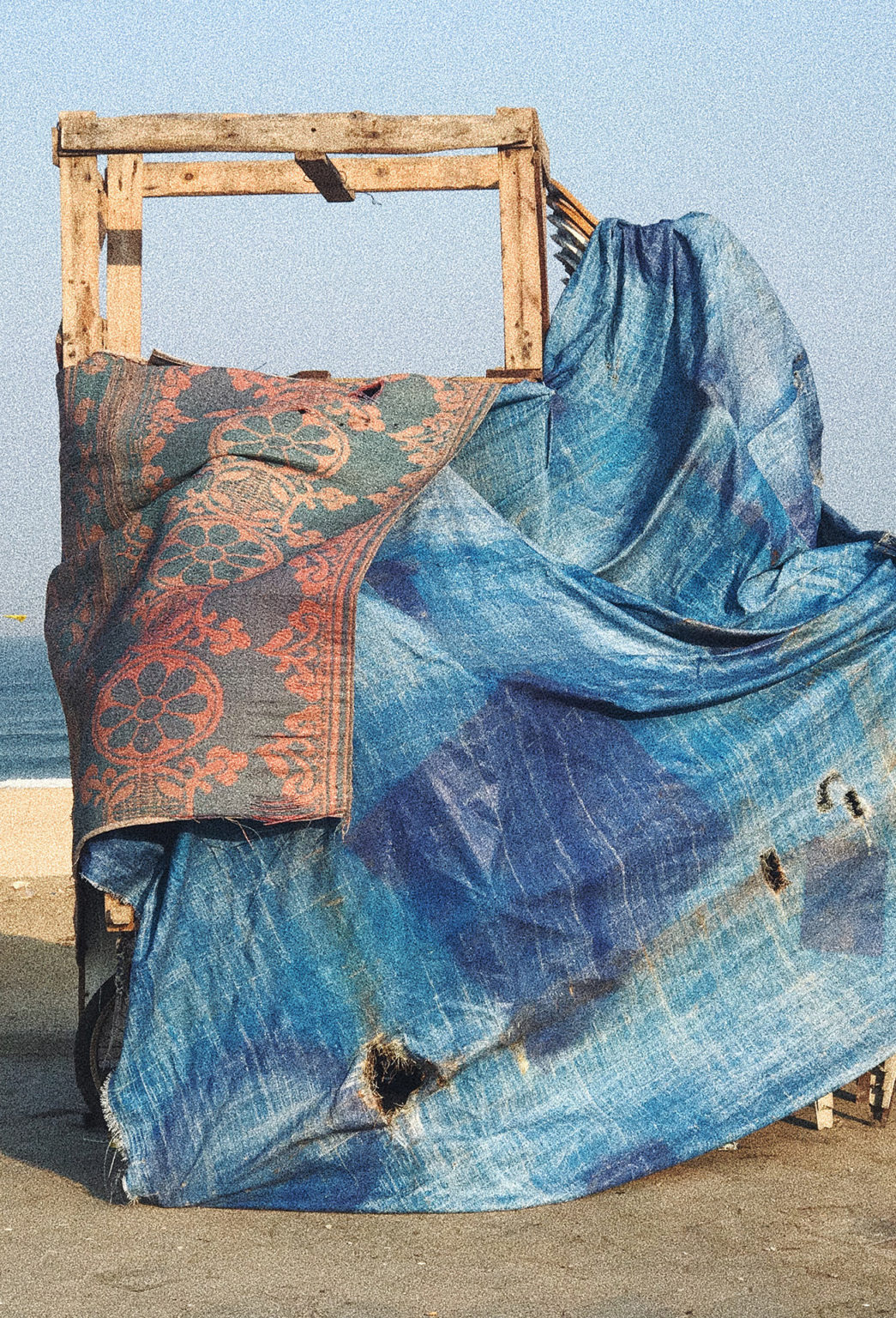 Photo d'une construction faite de planches de bois, et recouverte d'une bâche bleue délavée et trouée ainsi qu'un tapis de type oriental de couleur rouge et bleu se trouve sur le sable d'une plage. En second plan, on aperçoit la mer bleu.