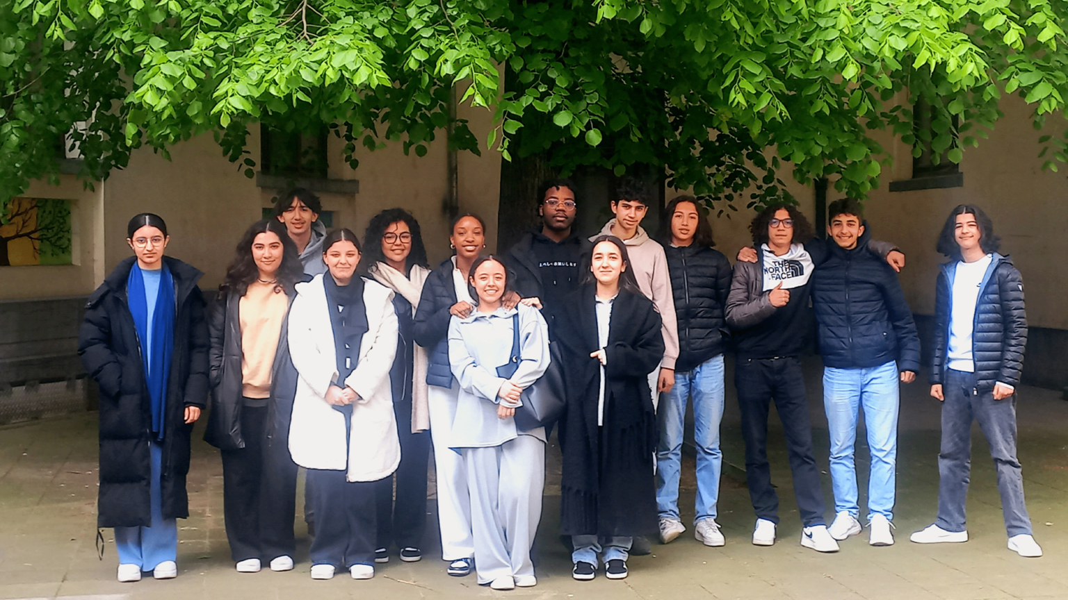 On retrouve une classe de jeune adolescent. Ils posent en groupe pour se faire prendre en photo, une photo de classe. Certains sont habillés en plus foncé tandis que d'autres sont en clair. Ils sont devant un bâtiment jaune et en dessous d'un arbre.