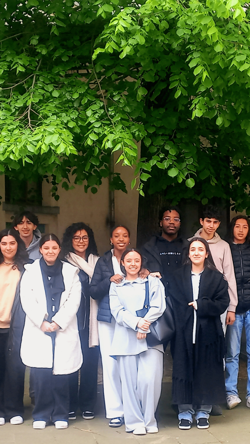 On retrouve une classe de jeune adolescent. Ils posent en groupe pour se faire prendre en photo, une photo de classe. Certains sont habillés en plus foncé tandis que d'autres sont en clair. Ils sont devant un bâtiment jaune et en dessous d'un arbre.
