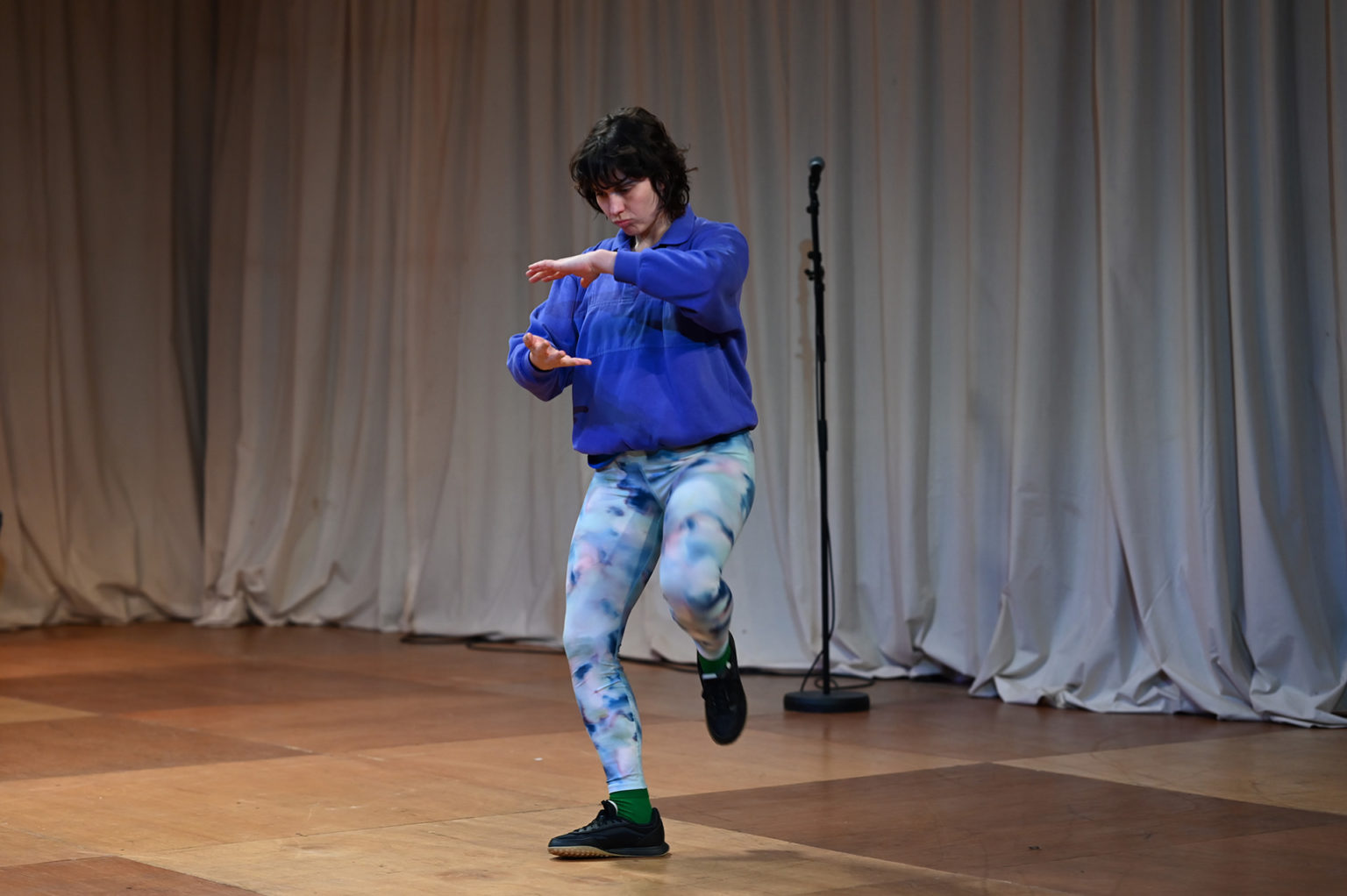 Chloé Larrère qui interprète Fani danse et fait un mouvement avec ses mains. Elle porte un legging et un pull bleu.