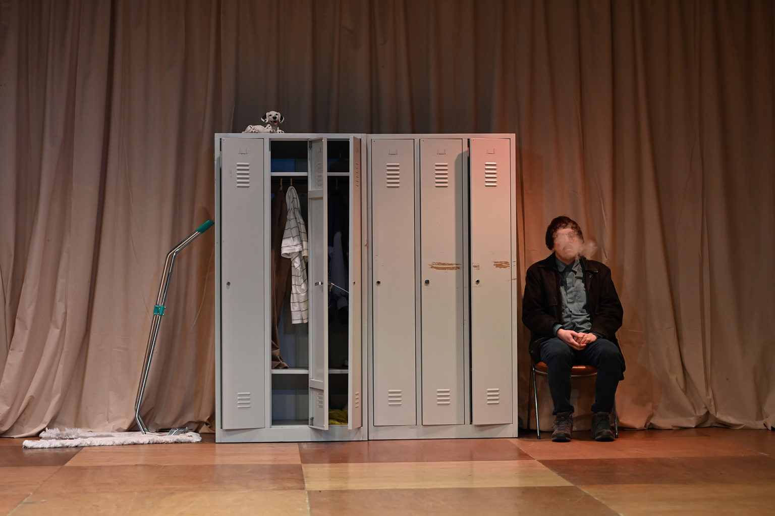Dans une salle, à côté d'une armoire à casiers, se trouve un comédien (Daniel Schmitz) qui joue le gardien de la salle. Il fume et est habillé de manière sombre.