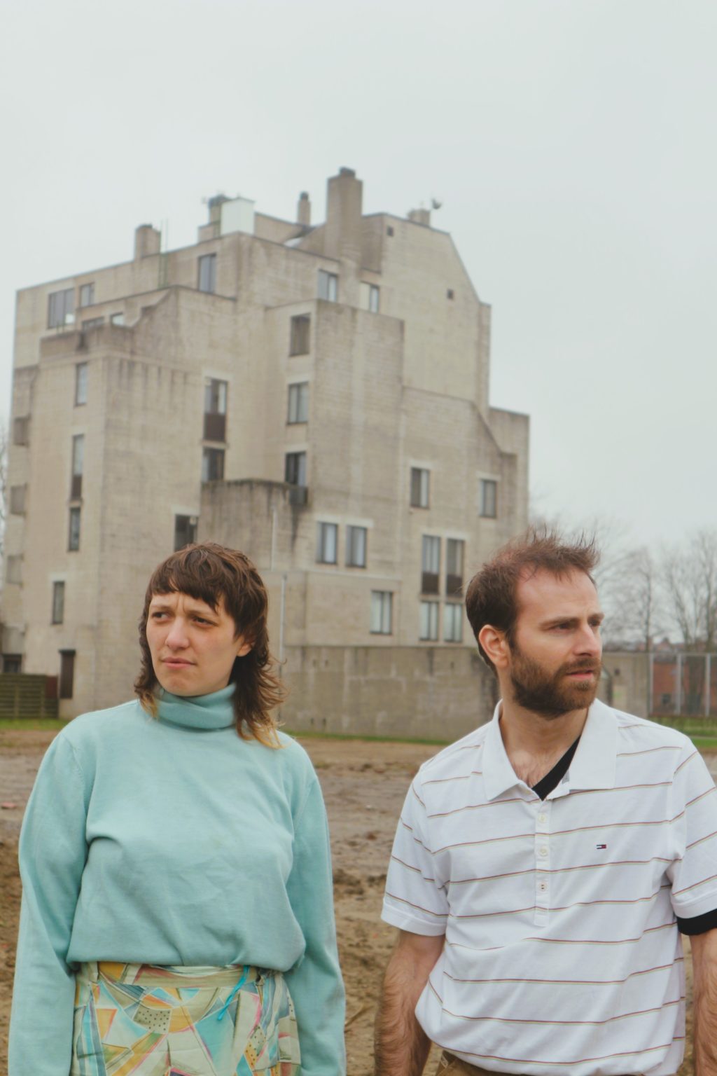 Photo de Cédric Coomans, Emilienne Flagothier devant un bâtiment. Cédric porte un t-shirt blanc avec des lignes noir et un pantalon beige. Emilienne porte un pull bleu clair et une jupe à motifs dans les mêmes tons.