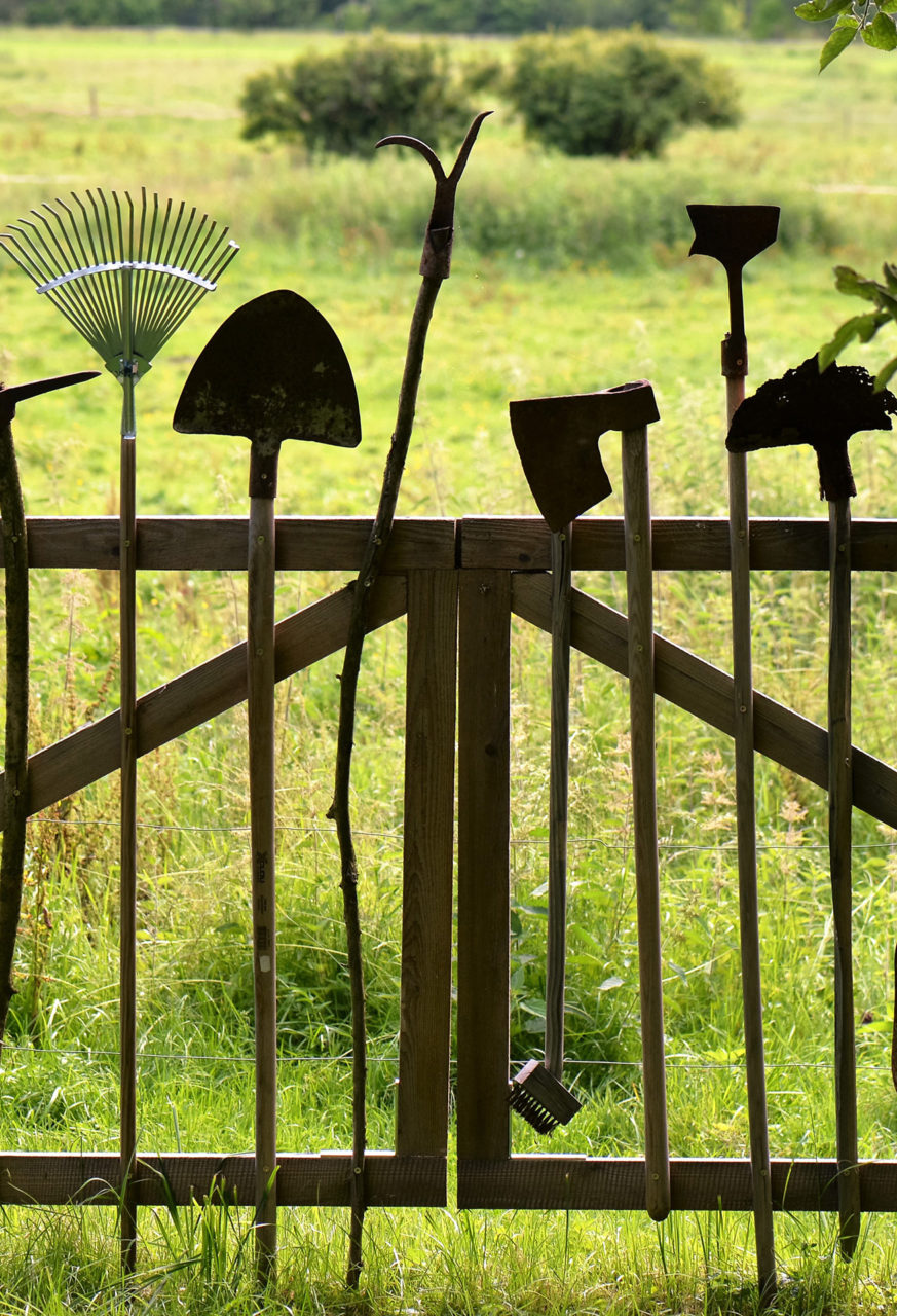 Une barrière faite d'outils de jardin est fermée et donne sur un grand pré.