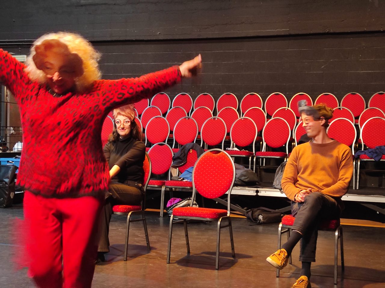 Photo d'un atelier théâtre sur le spectacle "L'amour c'est pour du beurre". Deux personnes sont assises, une autre est devant et lève les bras. Toutes portent un masque.