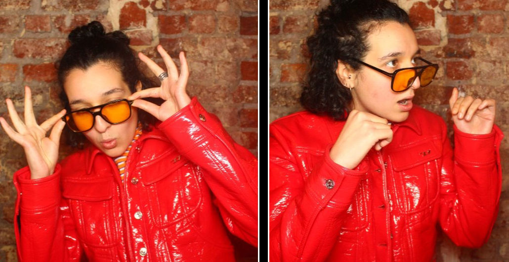 Syhame Youb, chargé de billetterie au Théâtre Les Tanneurs vêtue d'une veste rouge. Elle porte des lunettes de soleil style aviateur. Elle pose devant un mur en brique rouge.