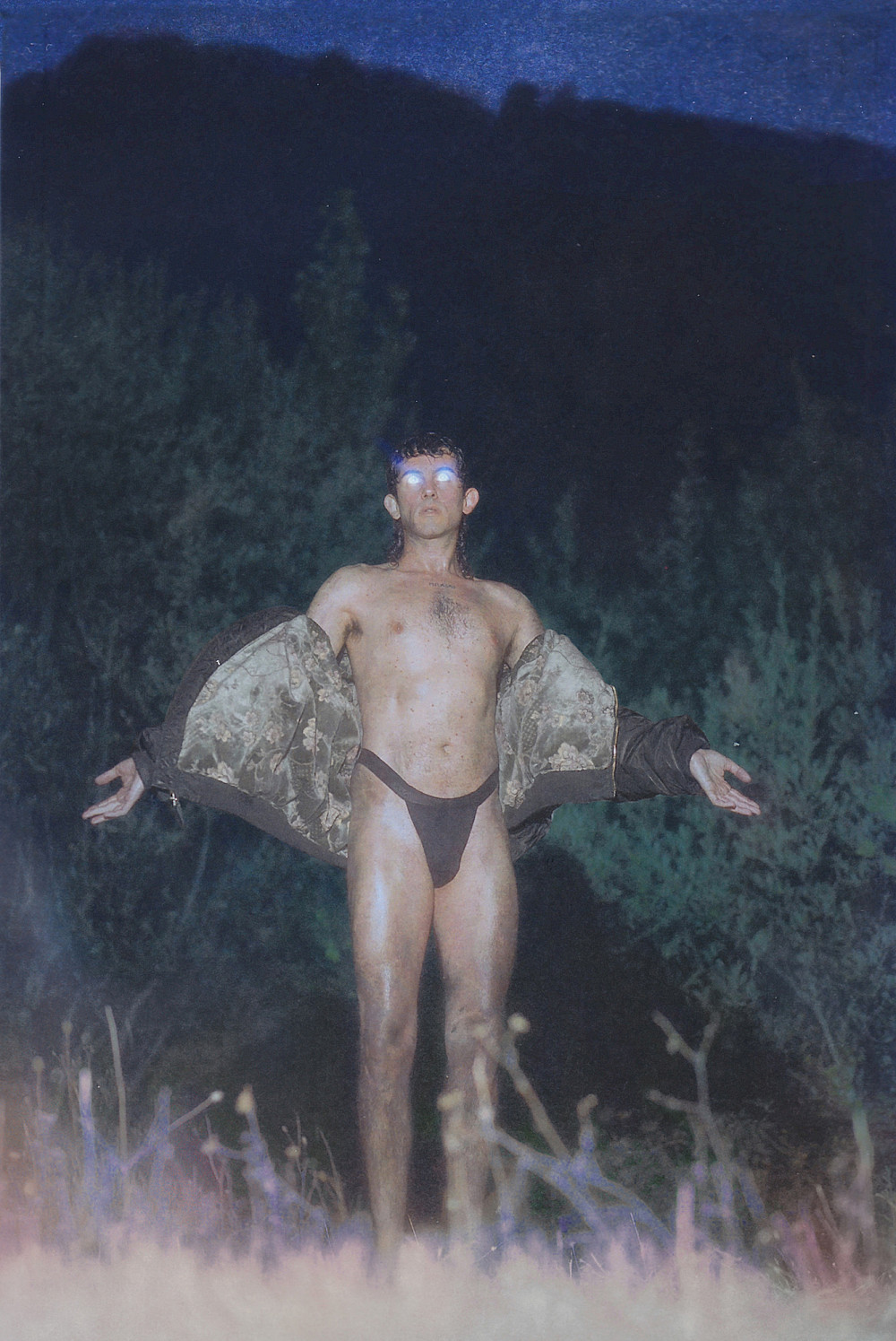 Alberto Cortés pose en sous-vêtement devant un champ à la tombée de la nuit. Il écarte les bars et ses yeux sont rempli d'une lumière blanche qui reflète.
