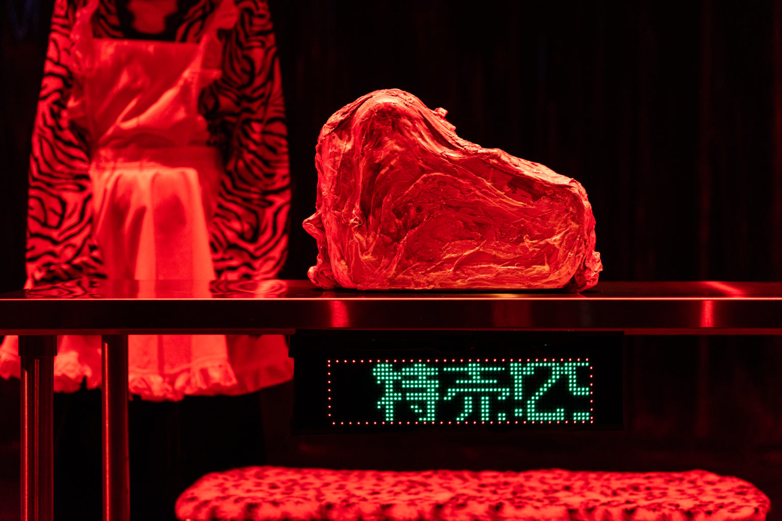 Un morceau de viande posée sur une table en acier en train de se faire pesé. Quelqu'un se tient derrière les bras le long du corps.