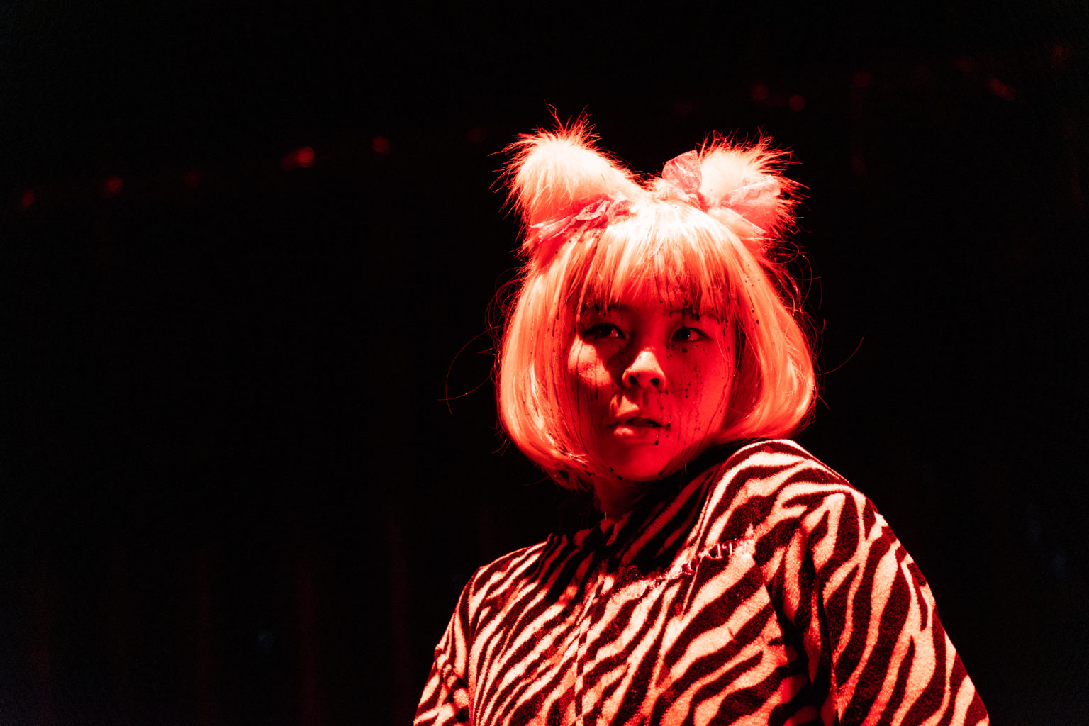 Une actrice asiatique avec des oreilles de chat posées sur ses cheveux blonds qui dévoilent une frange regarde au loin devant un fond noir.