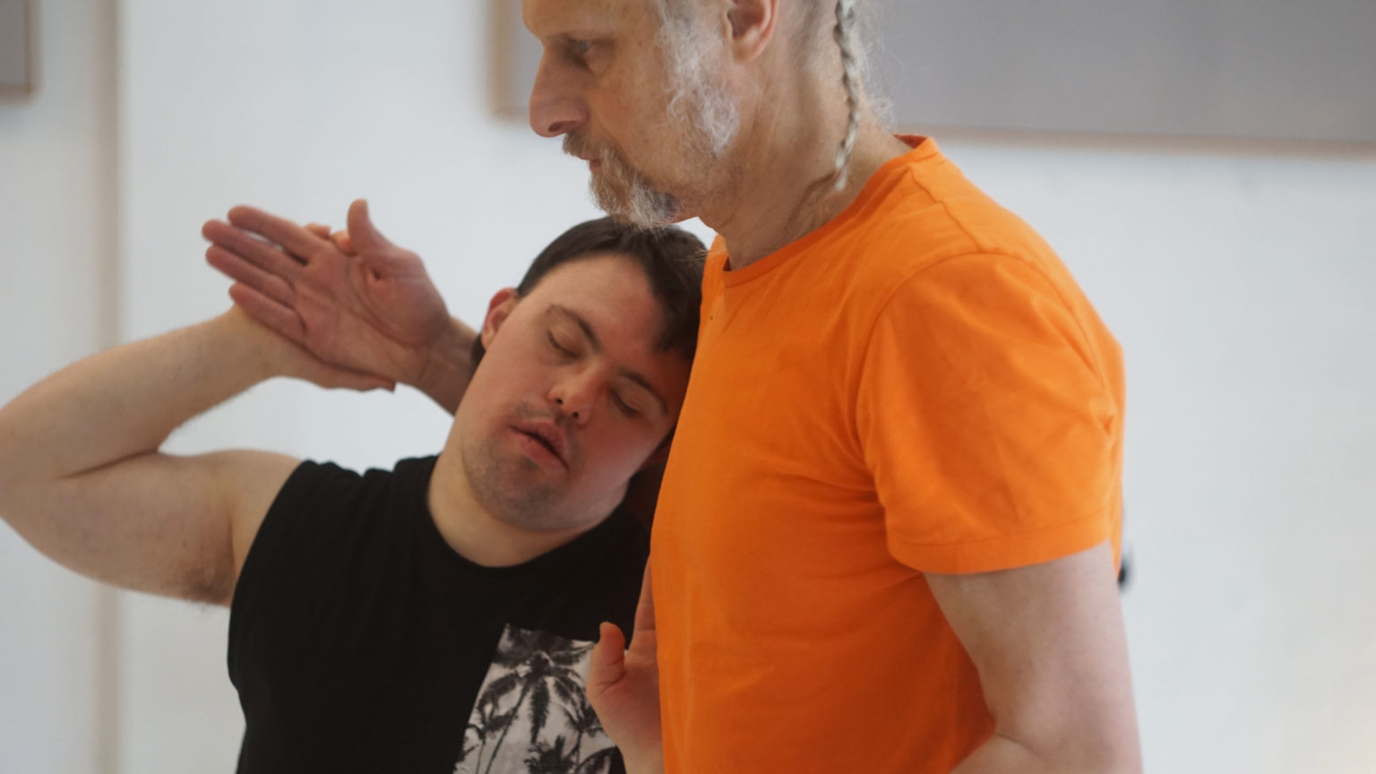 Un personne avec un t-shirt noir, les yeux fermés, repose sa tête sur la poitrine d'un autre homme avec un t-shirt orange et des cheveux gris. Ils sont en mouvement ensemble.