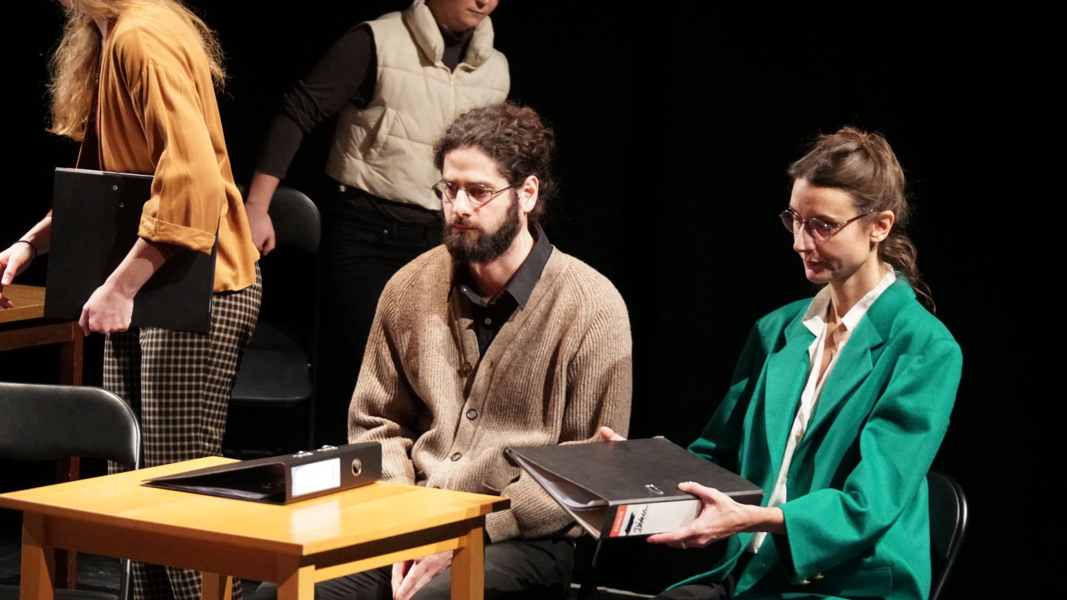 Un homme et une femme sont assis à une table avec un classeur posé dessus. La femme porte un blazer vert et des lunettes. Elle a les cheveux attachés et elle tient un classeur en main. L'homme porte des lunettes, une chemise noire et un cardigan marron. Il regarde le vide. Des gens sont en train de s'installer sur des chaises derrière elleux.