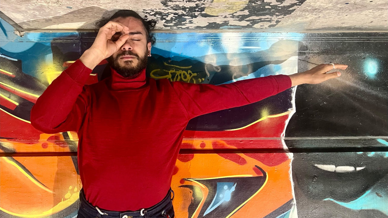 Mohamed Toukabri se tient devant un mur rempli de graffitis avec le bras tendu vers la droite et la main gauche en roind devant son oeil gauche. Il porte un pull rouge et à les cheveux attachés.
