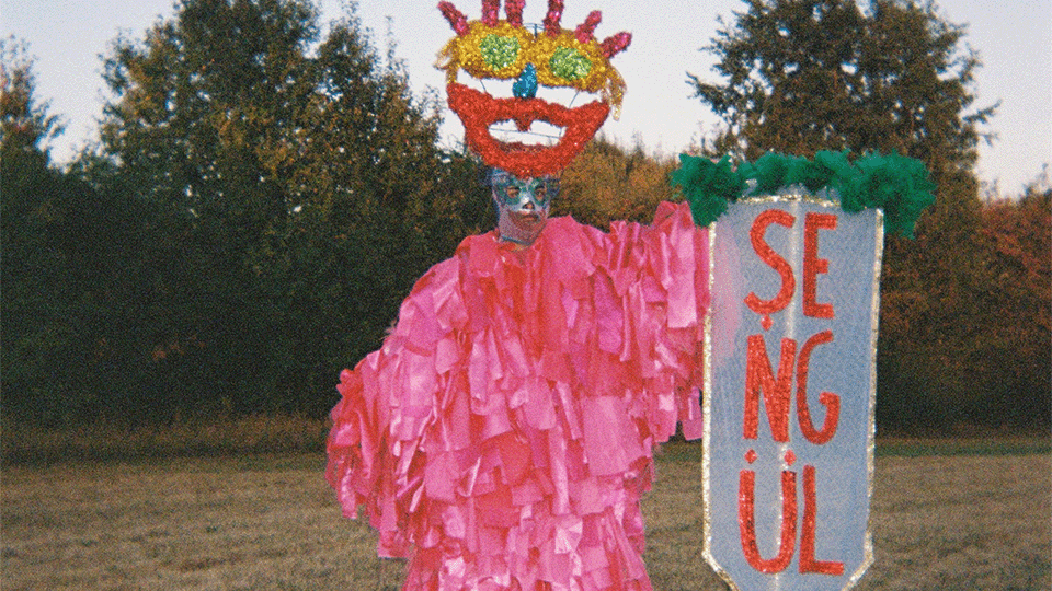 Une statue au visage coloré et déformé se tient au milieu d'une prairie, elle est vêtue de rubans roses et tient un étendard sur lequel est inscrit "Sengul"