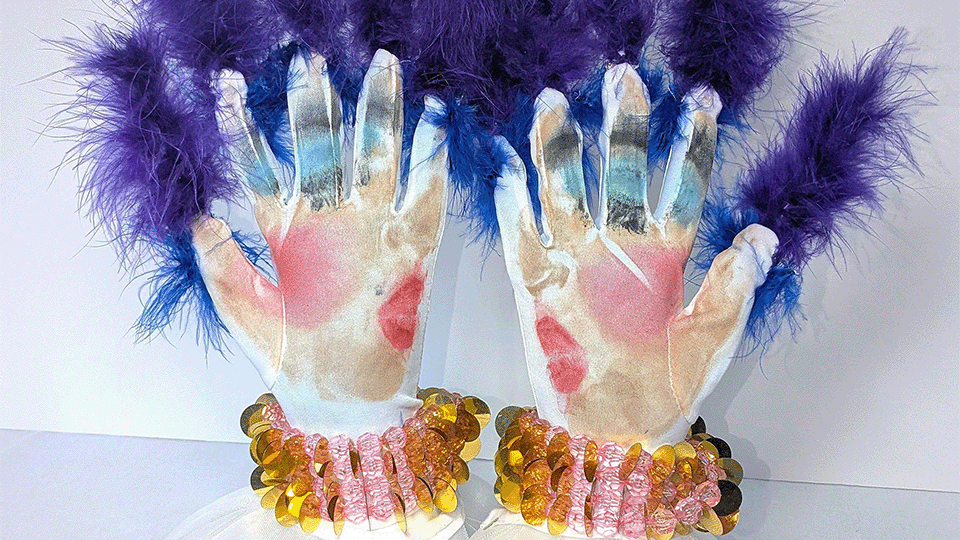 Deux gants sont exposés verticalement sur un meuble devant un mur blanc. Les gants sont colorés, présentant des plumes violettes aux bout des doigts et des bracelets dorés aux poignets.