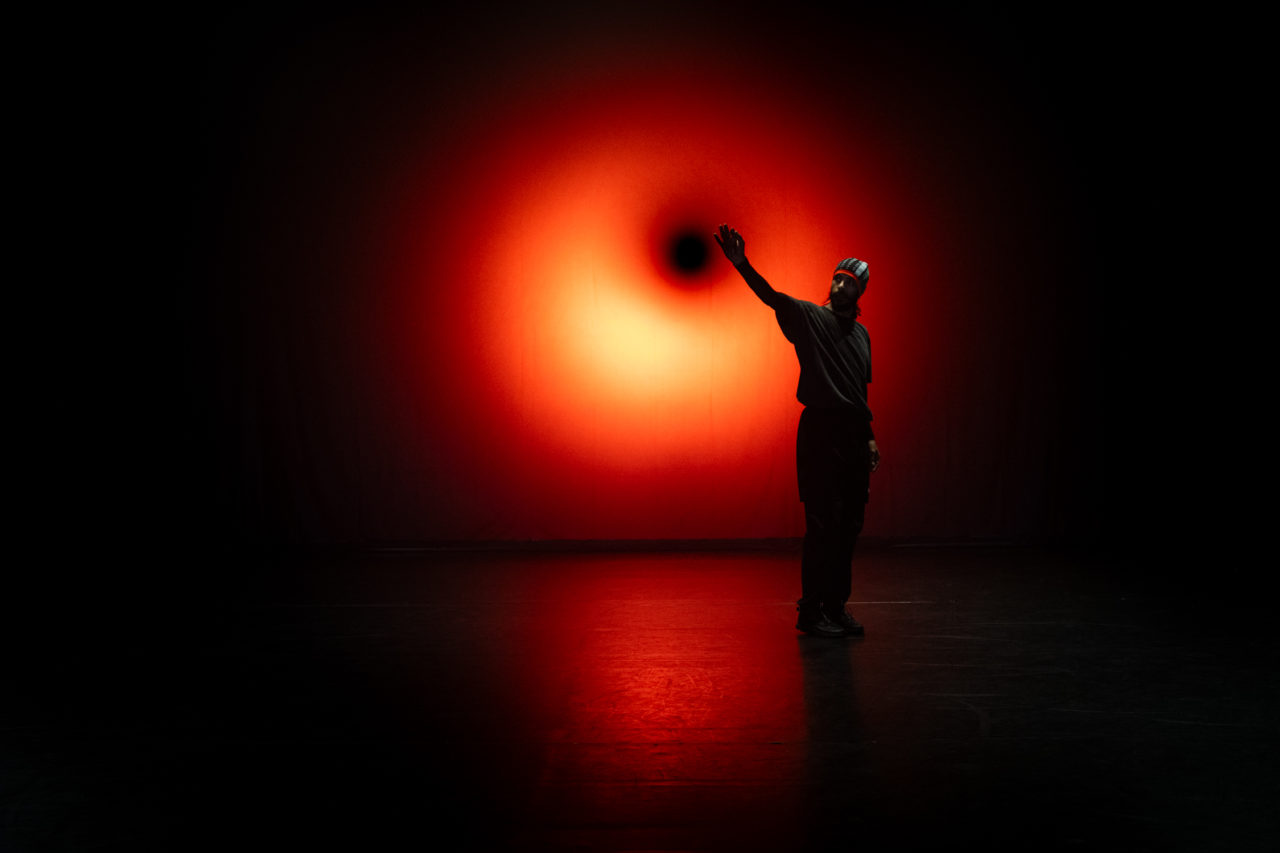 Mohamed Toukabri se tient debout, une main levée ver le haut. Il est à contre-jour, une lumière rouge orangée se trouvant derrière lui. On peut voir son visage.