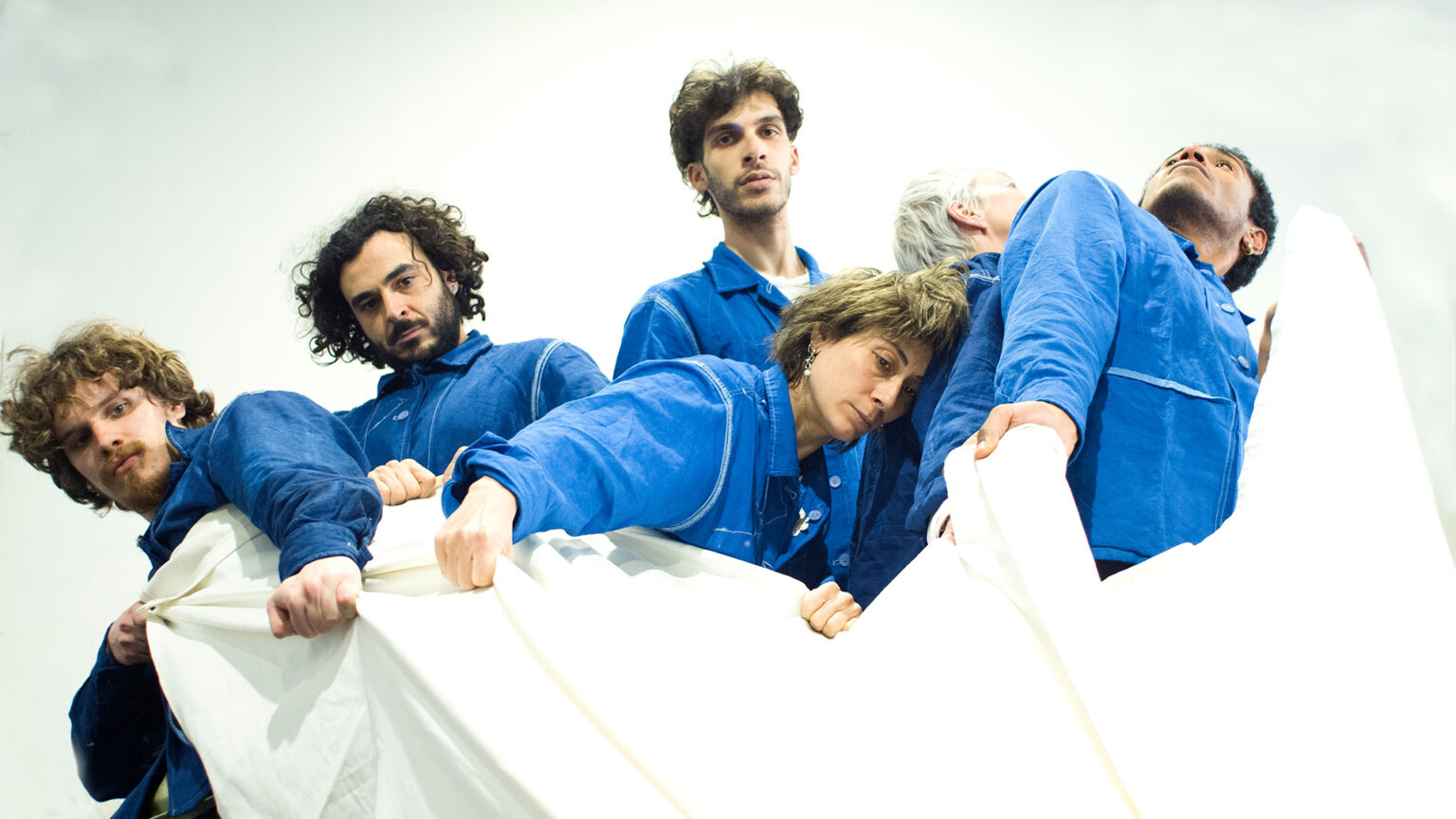 Groupe de personnes en combinaisons bleues tenant un grand tissu blanc. Les mains agrippent le tissu. Les corps sont serrés les uns contre les autres. Le fond est clair.
