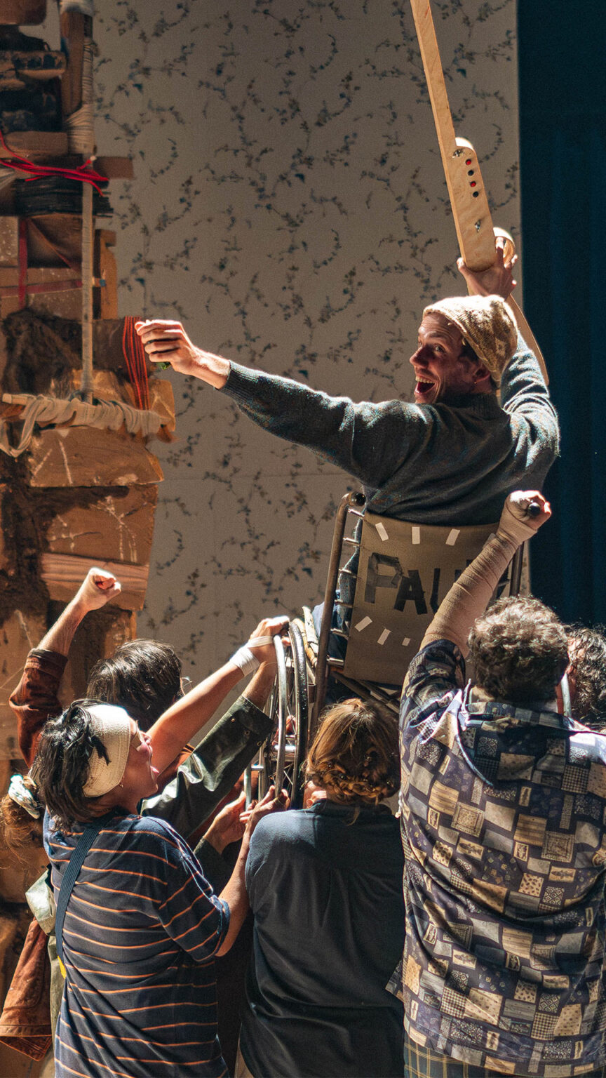 Scène de théâtre avec plusieurs personnes regroupées au pied d’une structure verticale faite de cartons et d’objets empilés. Une personne est en hauteur, appuyée sur l’installation. Des bras sont levés. Le décor comprend un mur clair à motifs et un rideau sombre sur le côté.