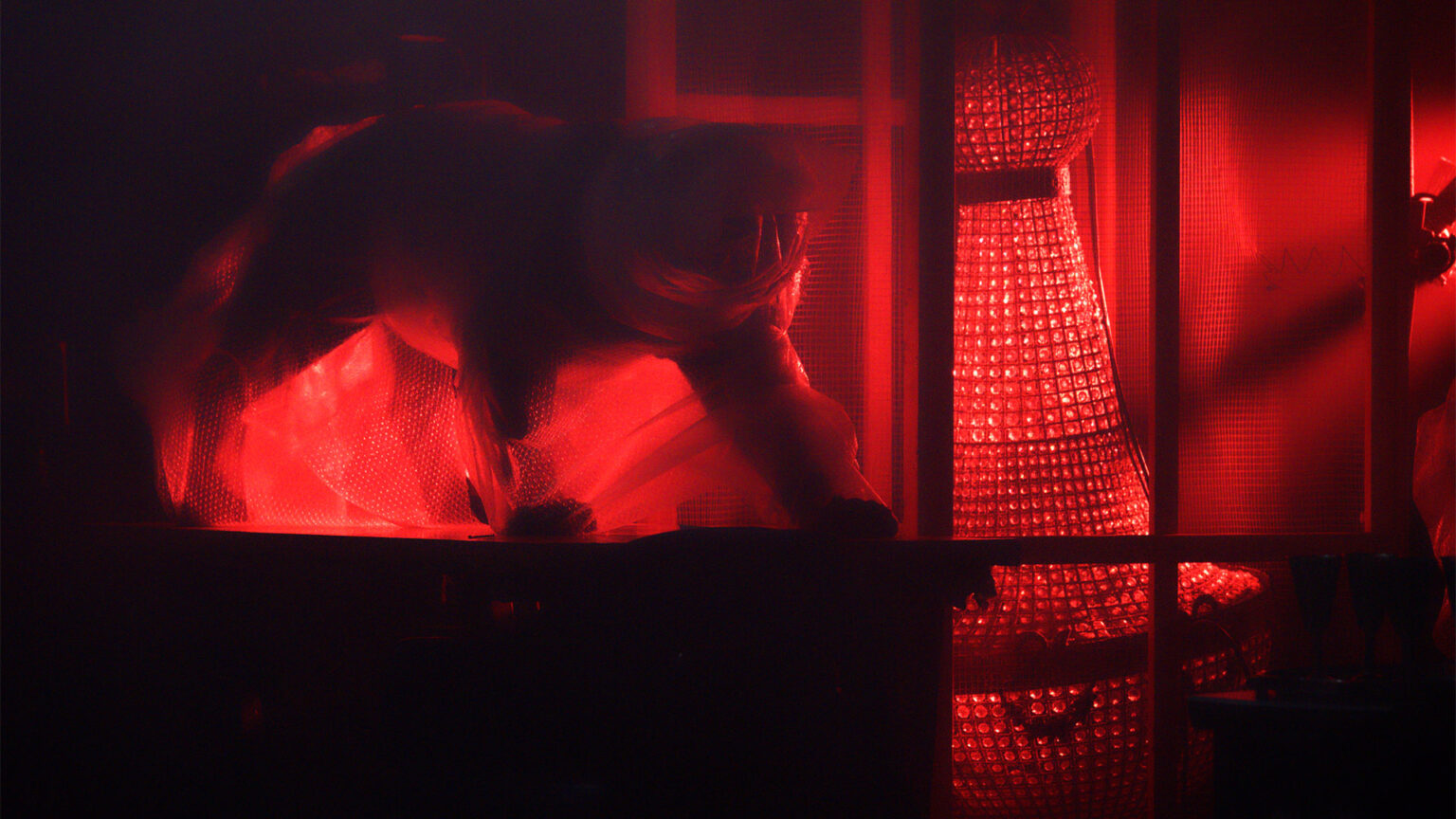 Scène sombre éclairée en rouge. Une personne est penchée vers l’avant, partiellement visible. Des éléments verticaux évoquant une grille occupent le premier plan. Un objet lumineux rouge se tient à droite. Le décor est fermé et fortement contrasté.