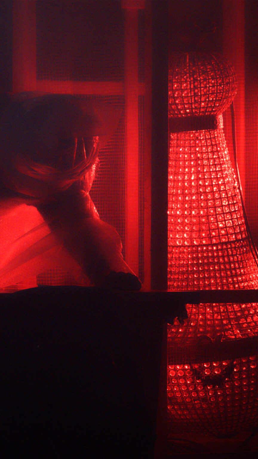Scène sombre éclairée en rouge. Une personne est penchée vers l’avant, partiellement visible. Des éléments verticaux évoquant une grille occupent le premier plan. Un objet lumineux rouge se tient à droite. Le décor est fermé et fortement contrasté.