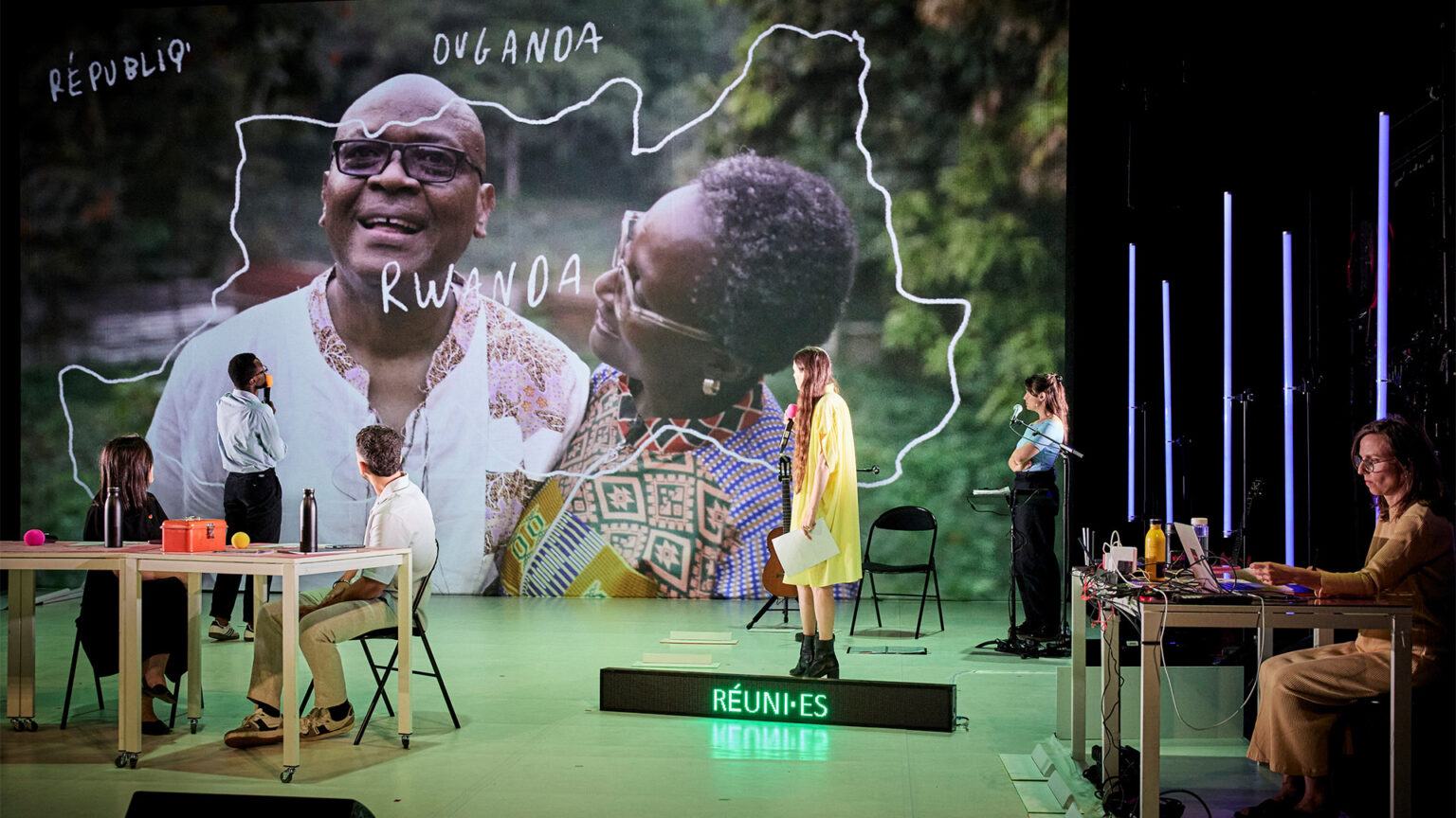 Plateau de théâtre avec plusieurs personnes réparties dans l’espace. En fond, une grande image projetée montre deux personnes devant un décor naturel. Des mots sont dessinés sur l’image, dont « RWANDA ». Des tables, des micros et un écran lumineux au sol sont visibles.