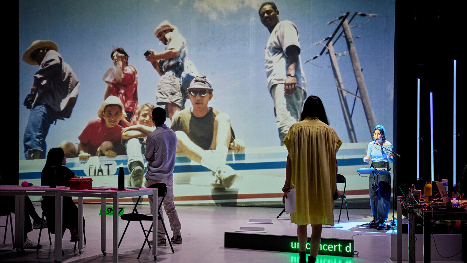 Une grande projection montre un groupe de personnes posant sur un bateau. Sur le plateau, plusieurs personnes sont debout ou assises à des tables. Des microphones et un pupitre sont présents. Un écran au sol affiche un texte lumineux partiellement visible.
