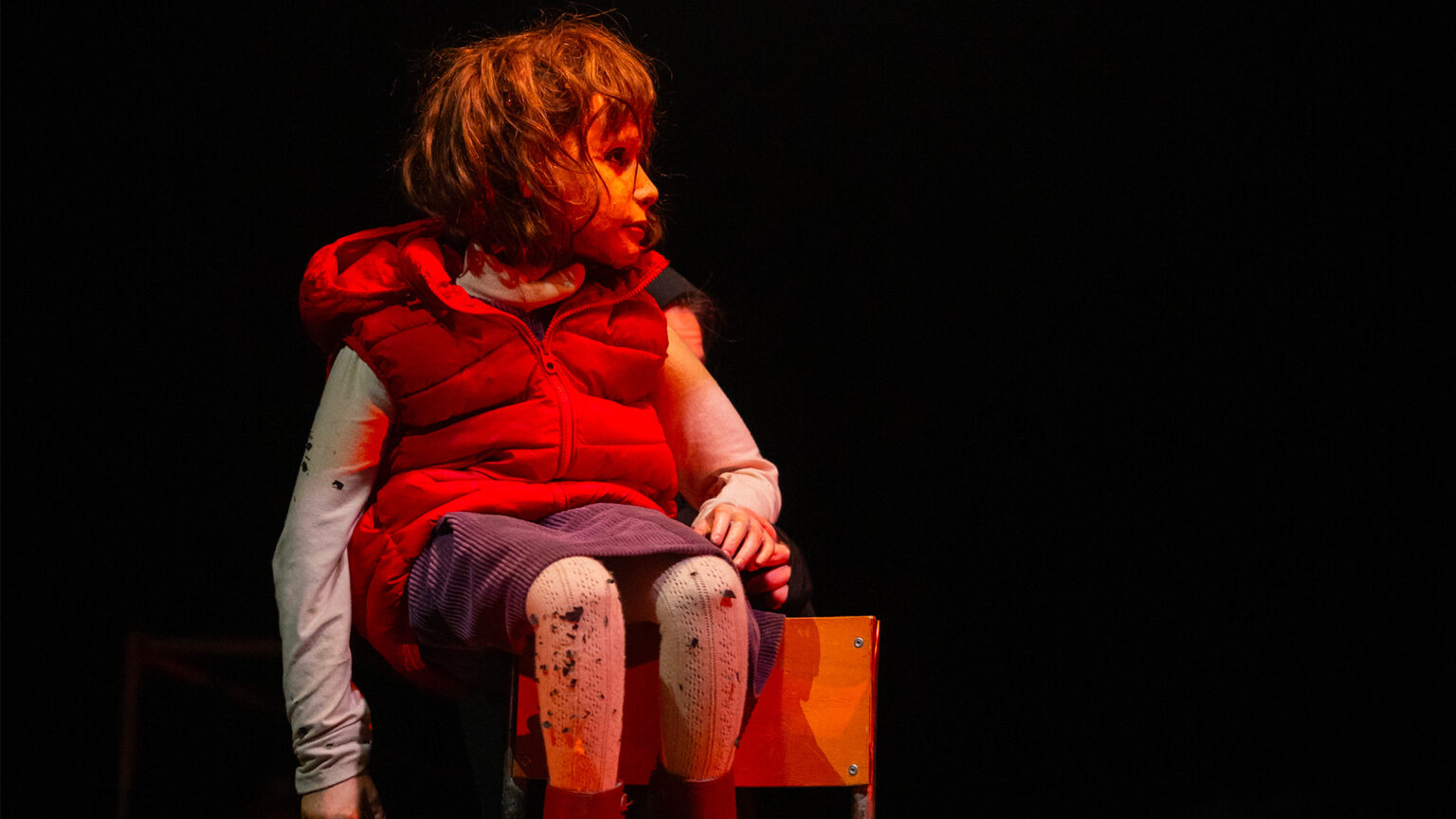 La photo montre une marionnette assise sur une petite caisse en bois, sur fond noir. La marionnette porte un gilet rouge, des vêtements clairs et a les jambes tachées de noir. Une actrice est visible partiellement derrière la marionnette, en arrière‑plan. L’éclairage met en valeur la marionnette, le reste de la scène est sombre.