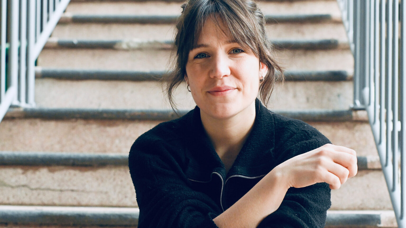 Portrait d’Héloïse Ravet assise sur un escalier. Le regard est dirigé vers l’objectif. Les mains sont visibles au premier plan. L’escalier forme des lignes horizontales en arrière-plan. Un garde-corps métallique est visible sur le côté.